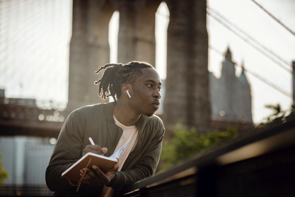 Black man writing notes in a notepad near a bridge outside