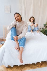 Upset Black woman sitting on the edge of the bed while her partner is preoccupied with his phone
