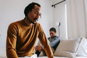 Black man sitting on the edge of the bed away from his sad female partner