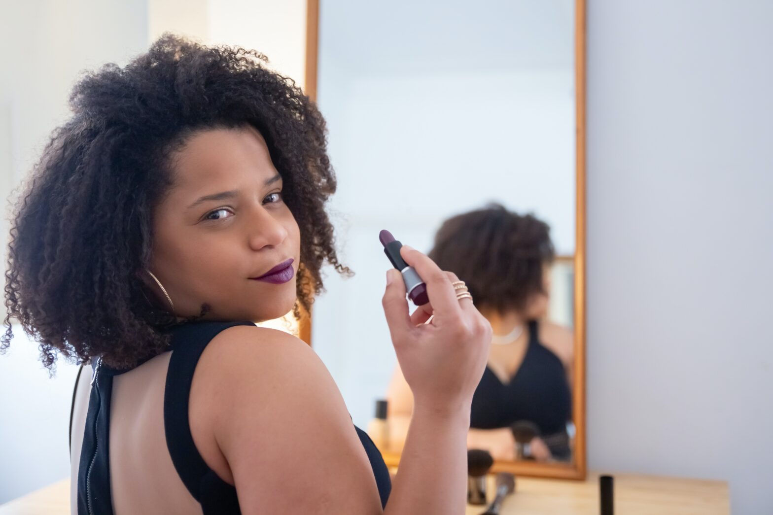 black woman in front of mirror looking at camera holding lipstick