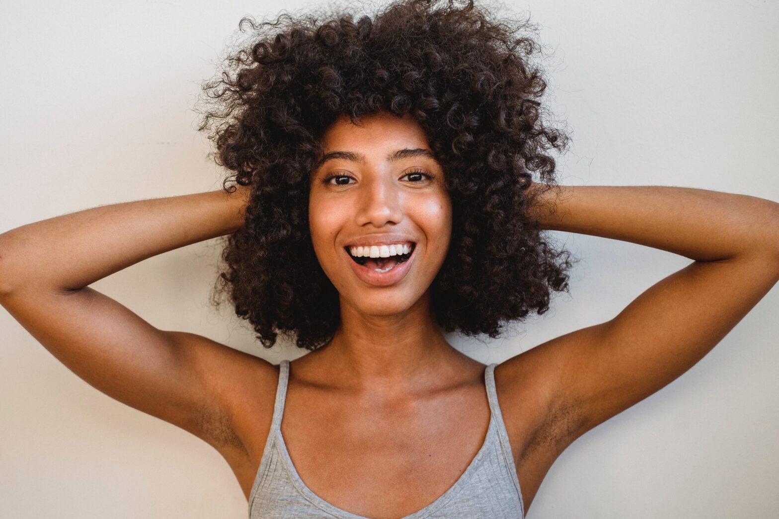 Black woman holds curly hair in grey tank top on grey background with an open mouth smile