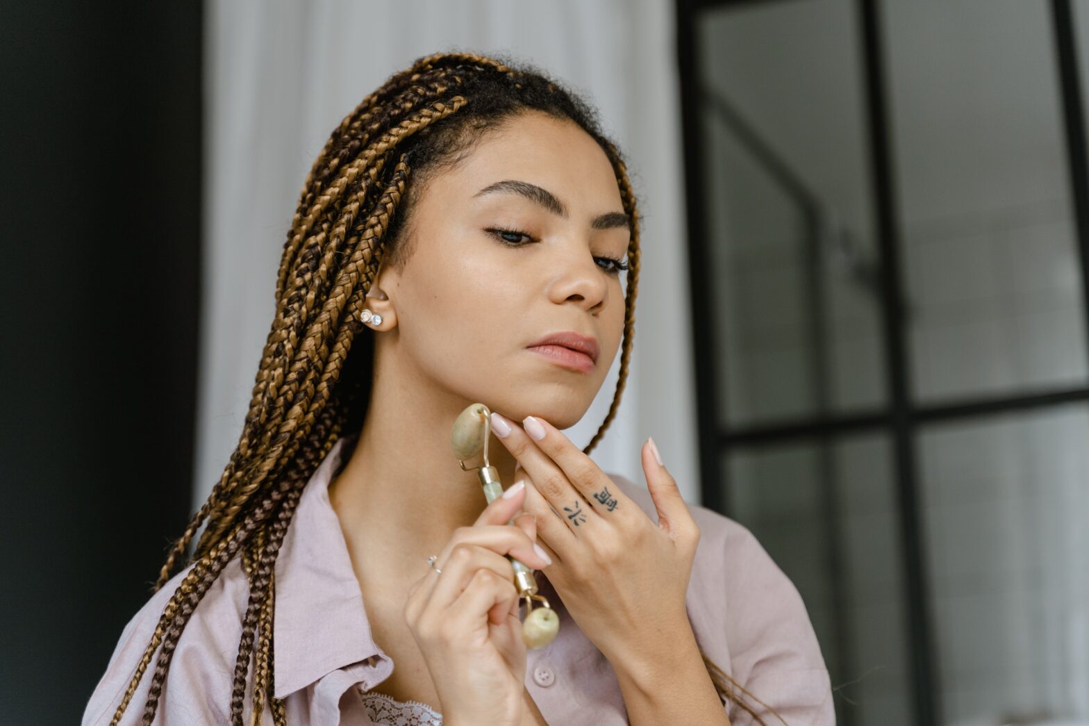 Black woman with knotless braids wearing pink pajamas using jade roller on her face