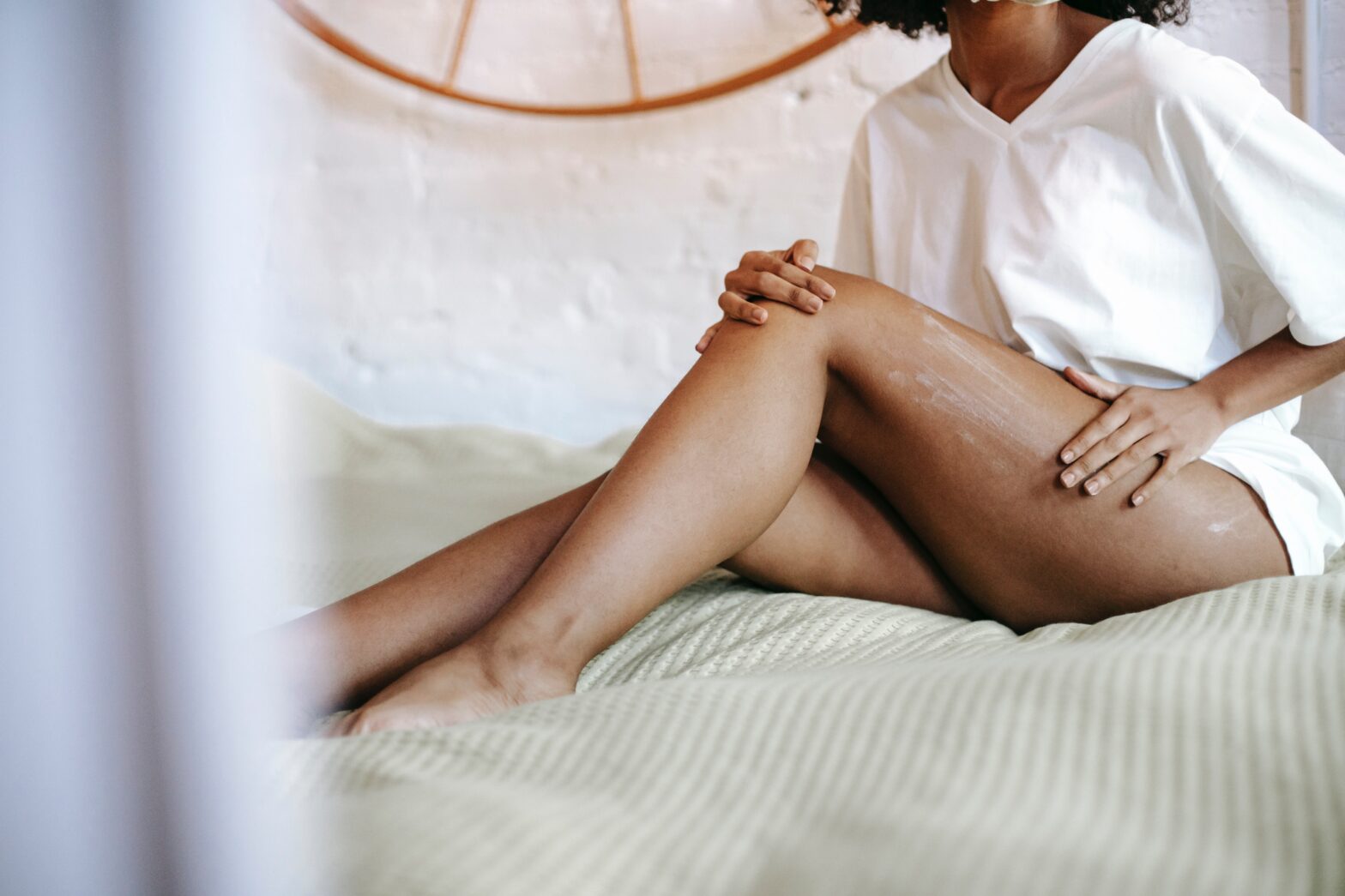Black woman massaging lotion into skin wearing a white shirt on plush bed