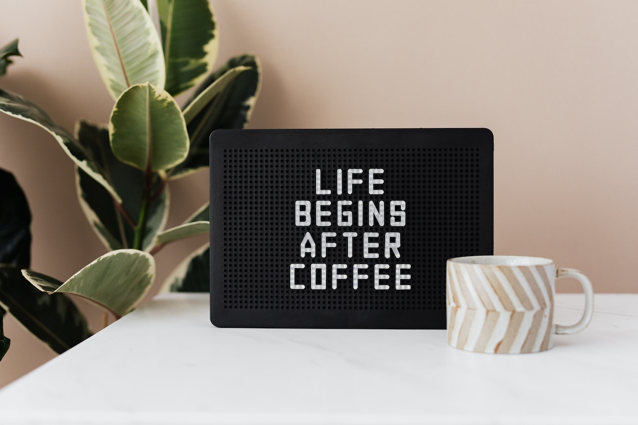 100 funny inspirational quotes to start your day off right with a laugh. Pictured: a note board that reads 'life begins after coffee' sitting on a white table next to a coffee mug and a house plant.