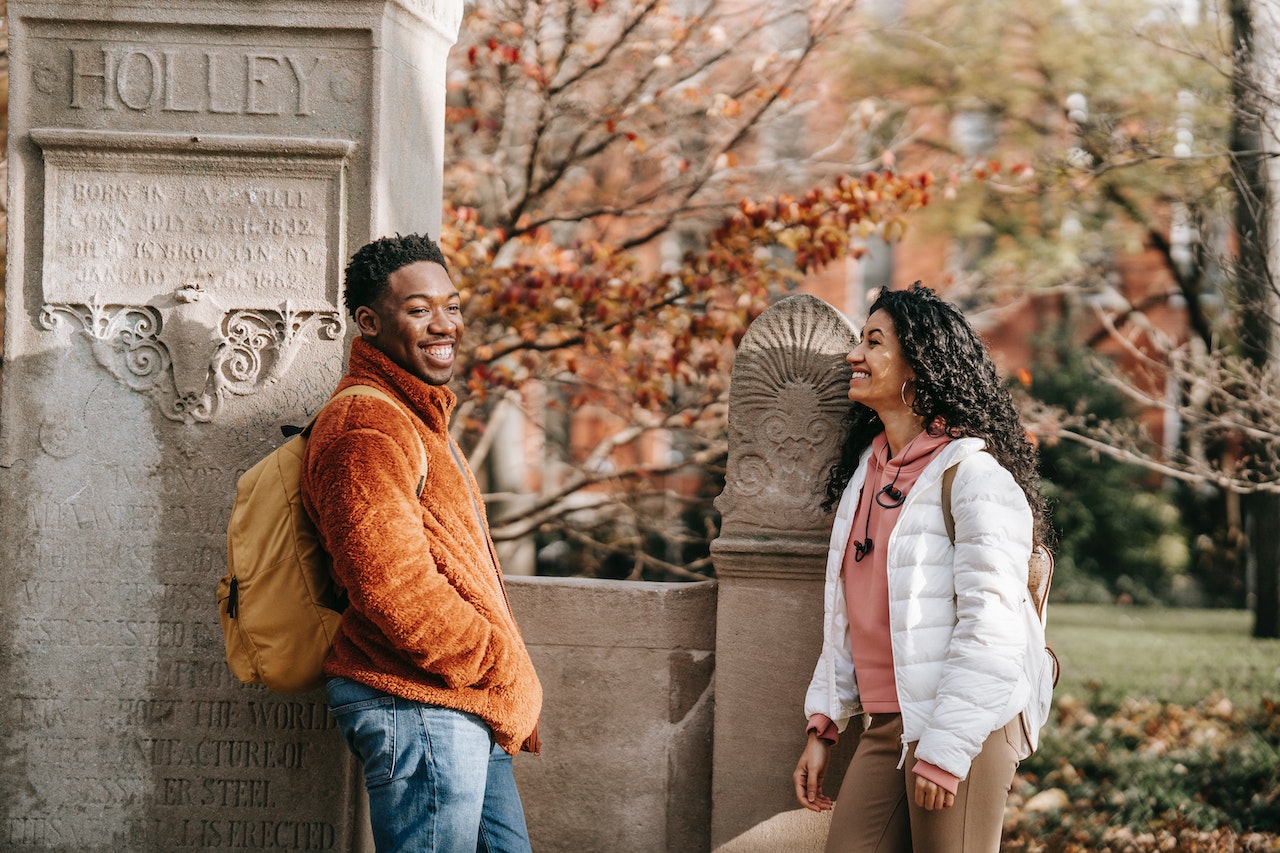 Make your first date filled with laughter with these 100 corny pick up lines. Pictured: A male student and a female student with backpacks standing in front of a monument while smiling at each other on a college campus.