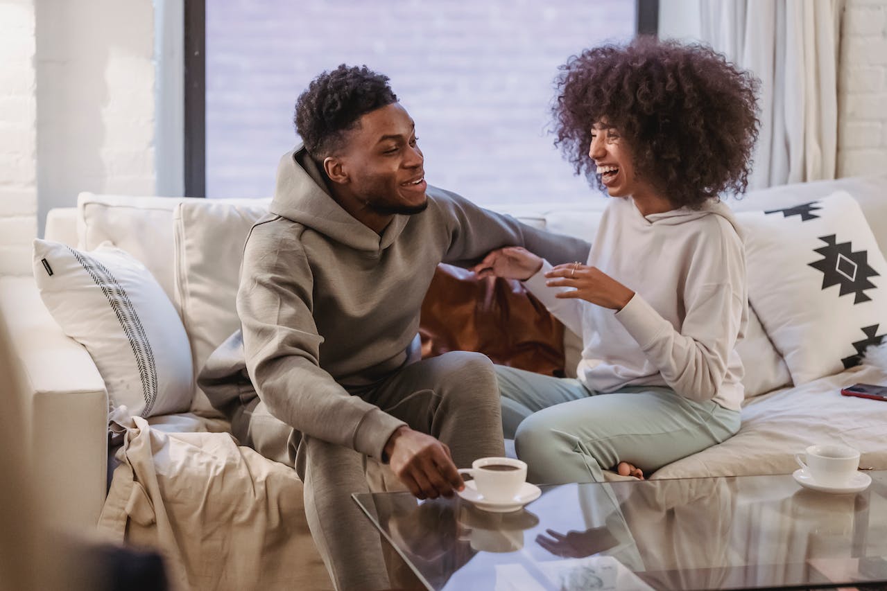 Black couple sitting on couch laughing together. These 25 romantic questions to get your boyfriend to laugh may be what your relationship needs.