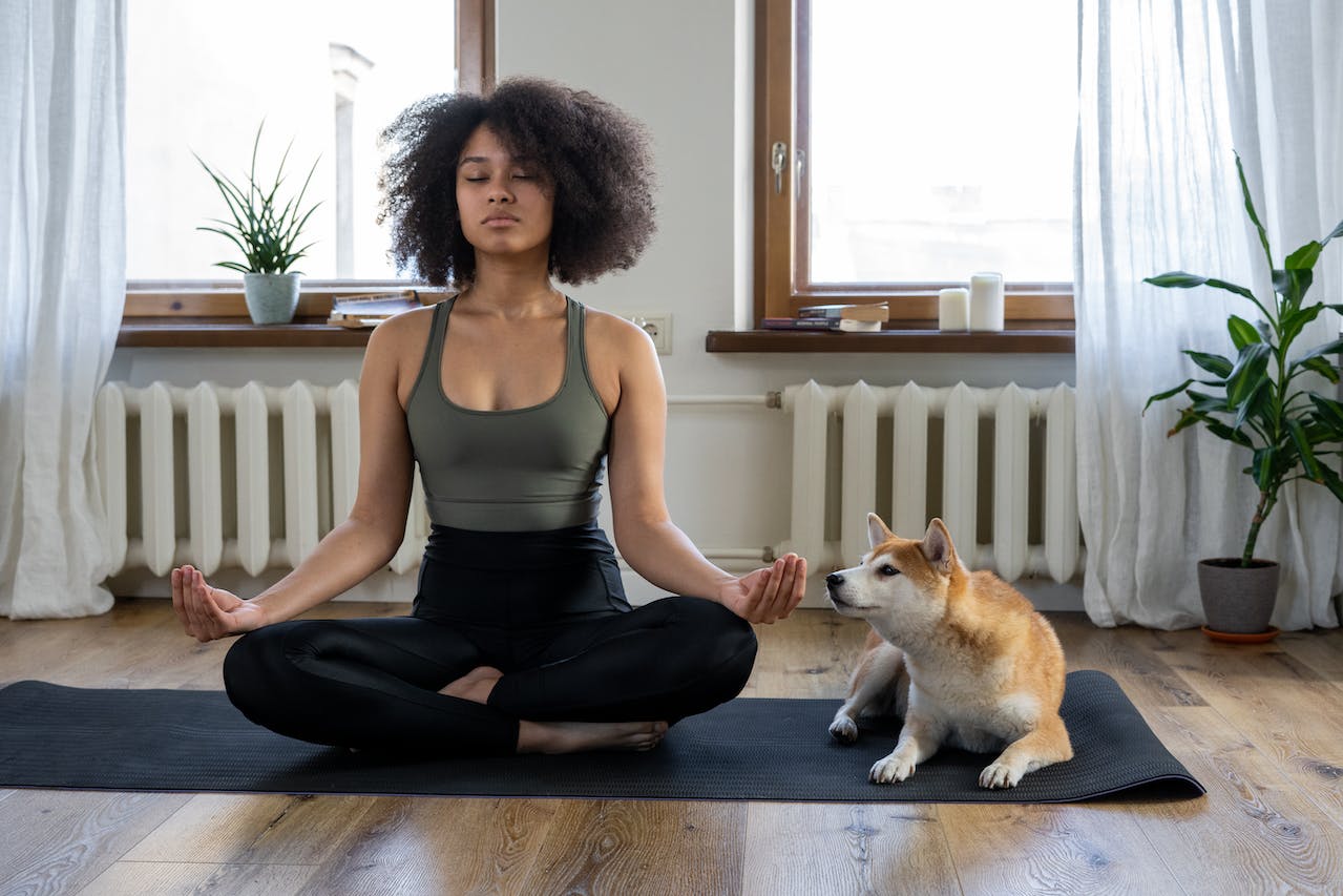 Change your perspective on life once you start enjoying its simple pleasures. Pictured: a woman meditating on a black mat with a dog trying to sniff her hand lying next to her.