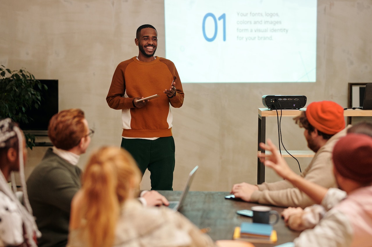Here are some PowerPoint night ideas that will make hilarious presentations to give to friends and family. Pictured: A man standing in front of a projector and giving a presentation to five other people.