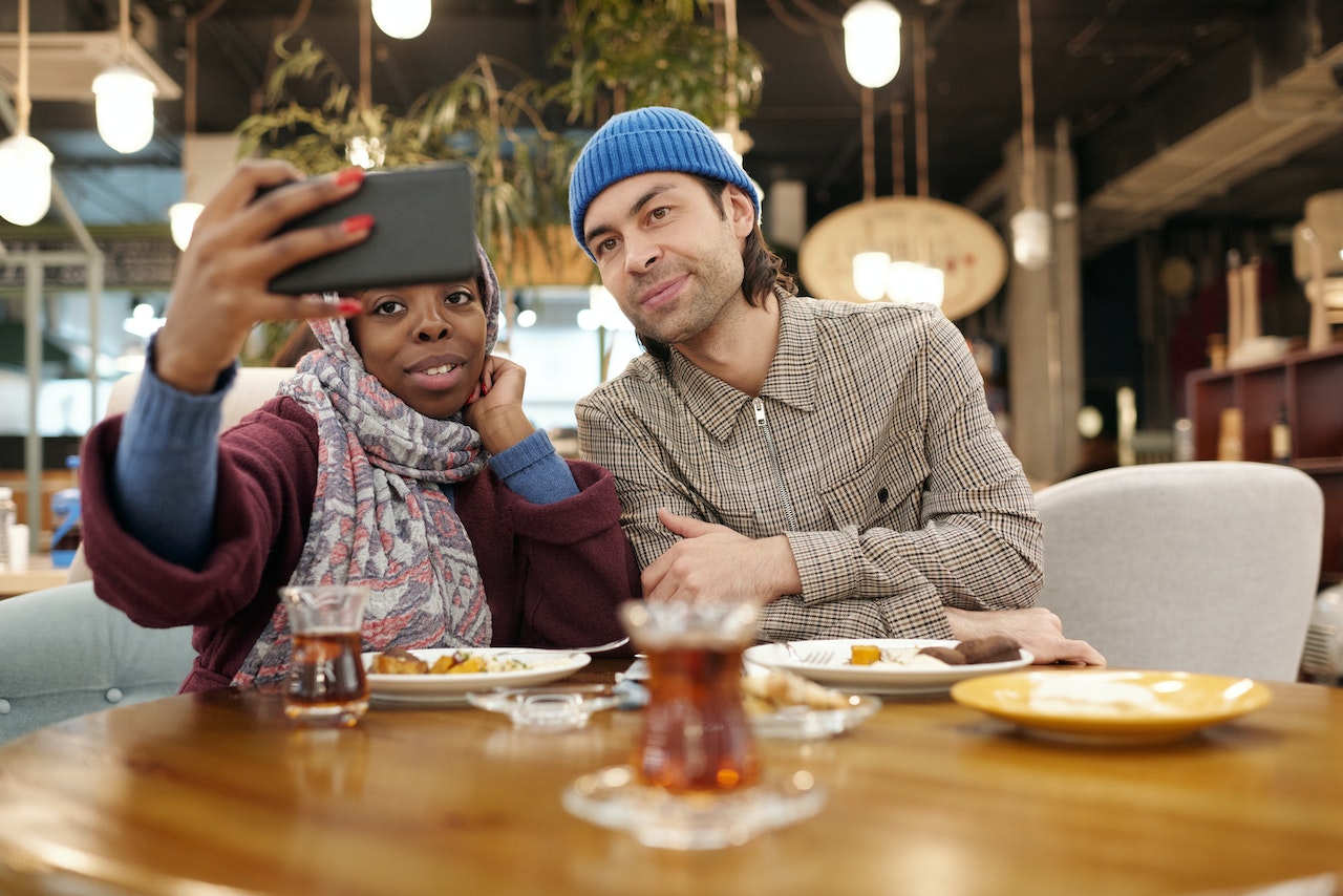 Is the olive theory of opposites attract a truism? Pictured: A woman holding a phone and taking a picture with a man while they sit in front of food in a restaurant.