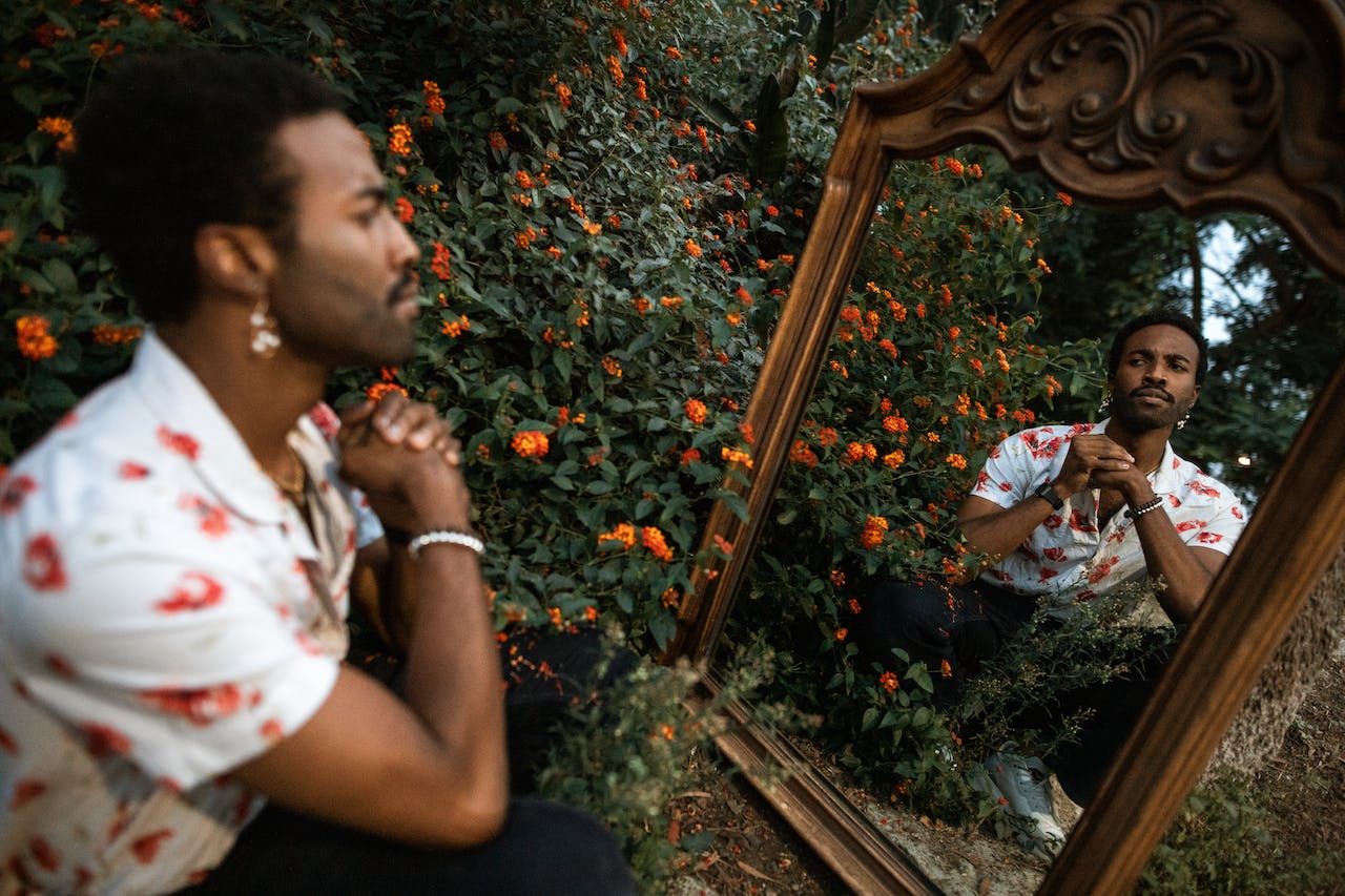 Here are 12 traits of a narcissist that can help you uncover whether you have one in your life. Pictured: a man kneeling in front of a mirror that is surrounded by orange flowers looking at his reflection.