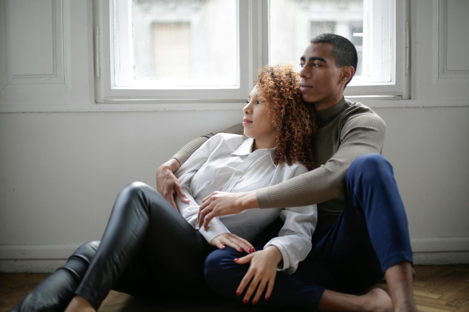 Thoughtful, young couple hugging while sitting on the floor near a window on a self-care date