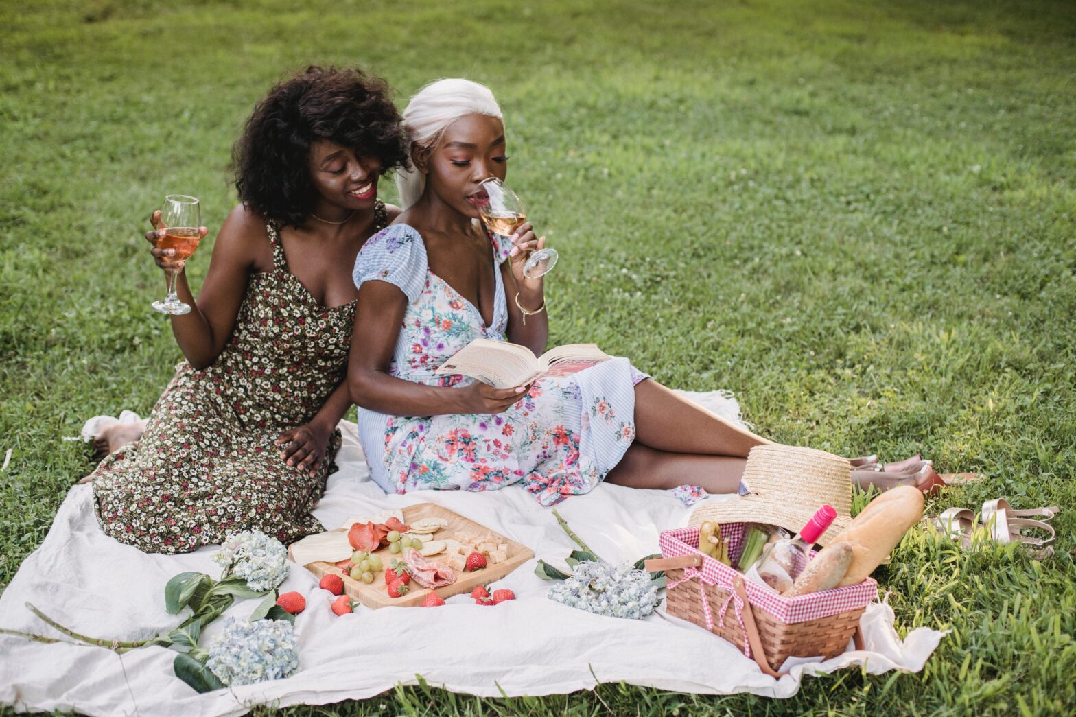 Two Black women in a park reading a book drinking wine picnic blanket dresses