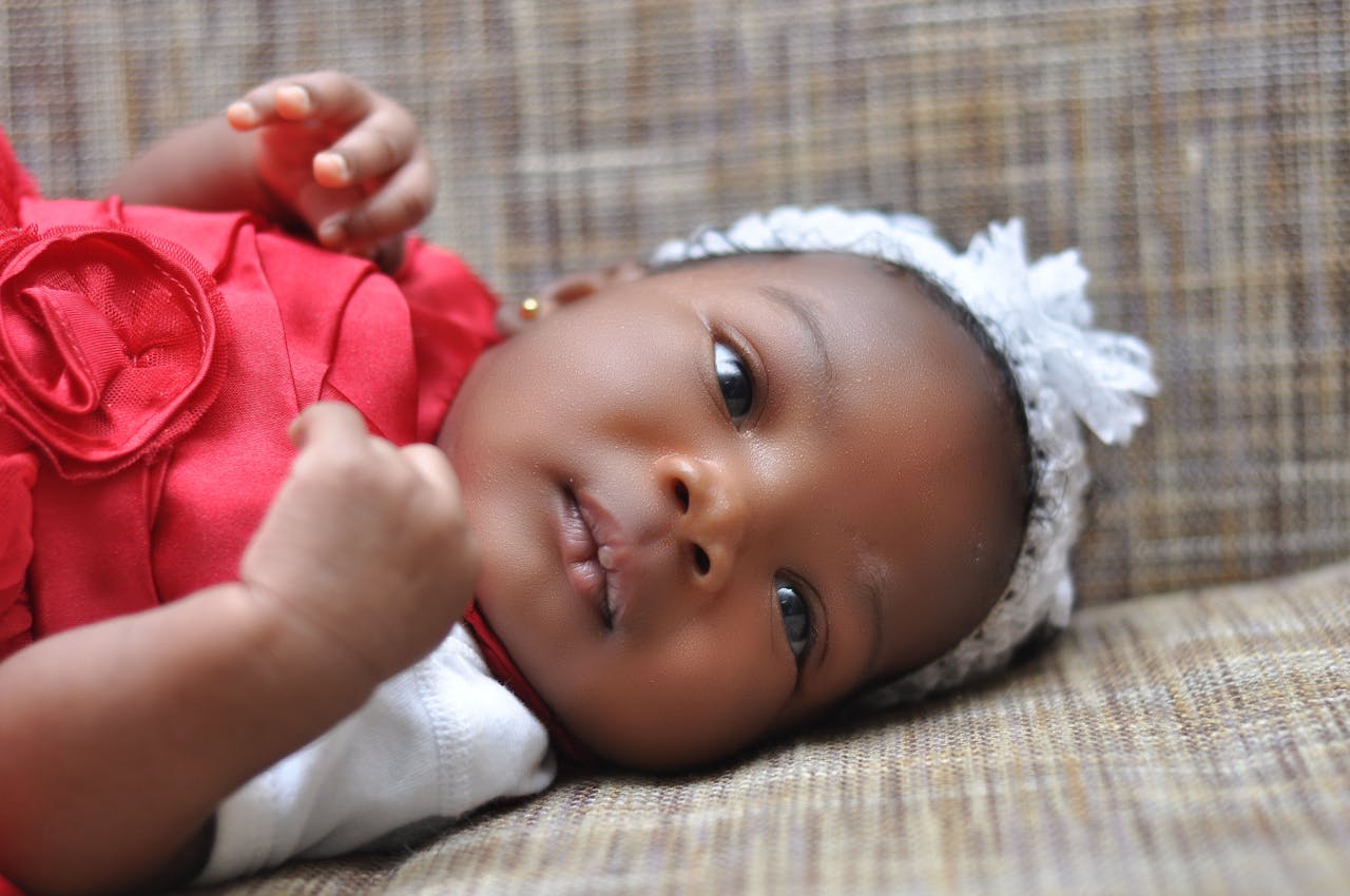 54 beautiful baby girl names that mean love. Pictured: a baby girl wearing a red dress and white head band lying down.