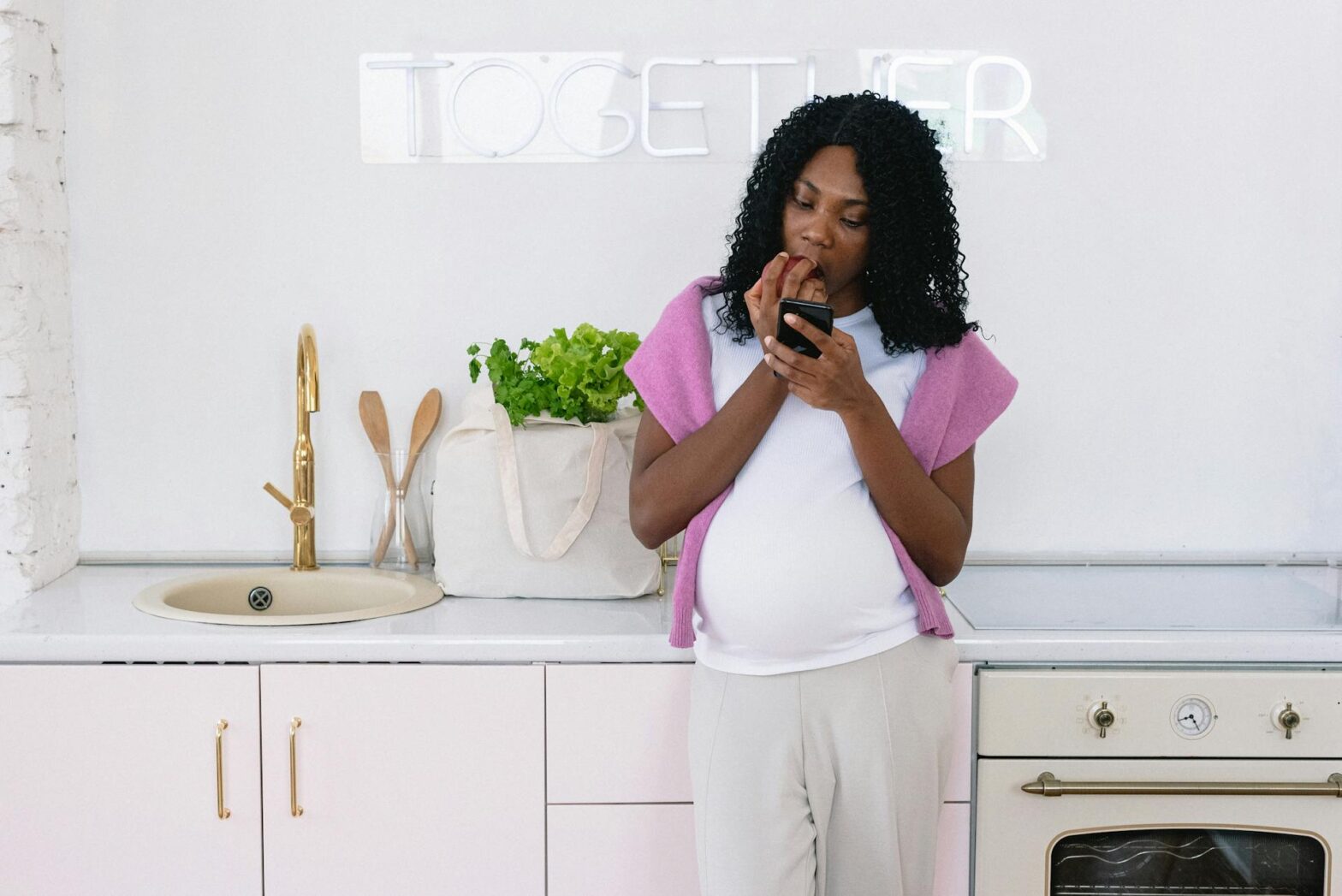 Black pregnant woman with smartphone eating in kitchen