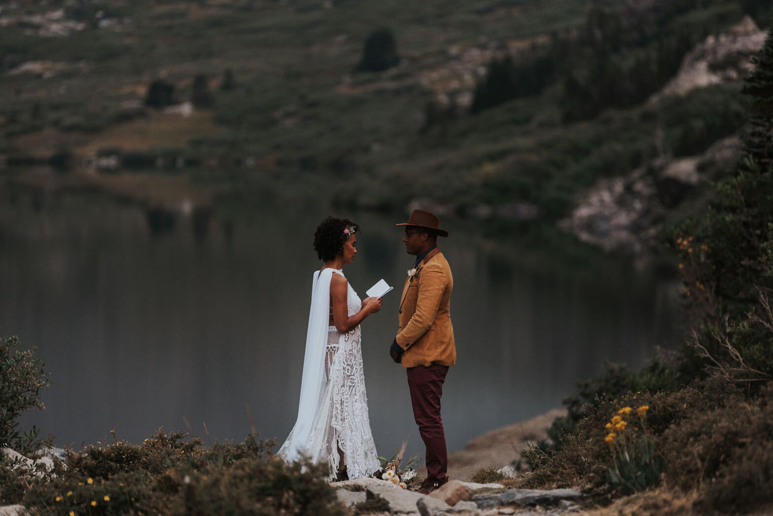 a bride with a cape wedding dress and groom with untraditional suit standing by mountains and water during elopement