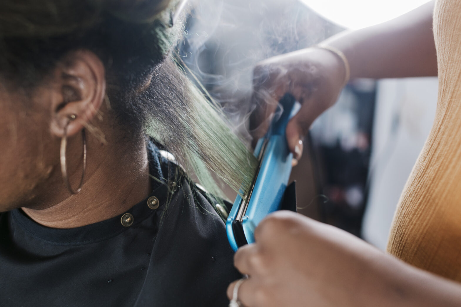 A close up of a black hairdresser straightening a client's hair with some steam rising from the heat.