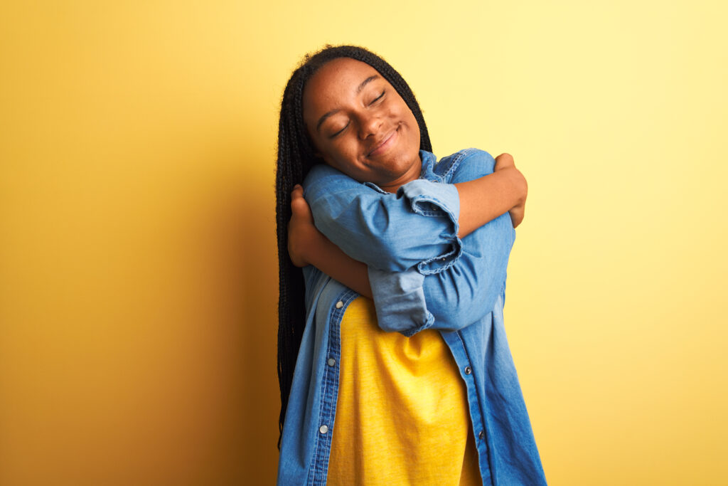 rejection therapy. Young african american woman wearing denim shirt standing over isolated yellow background Hugging oneself happy and positive, smiling confident. Self love and self care