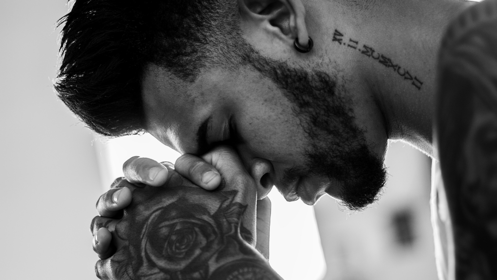 close up of a tattooed man resting his head on his folding hands roman numerals on neck and rose on hand