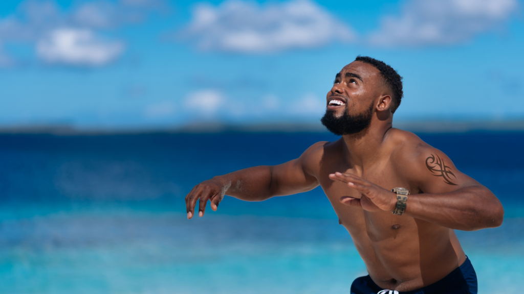 Black male playing volleyball on beach wearing watch and tribal tattoo on arm  with water in background