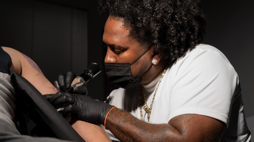 Male tattoo artist actively tattooing a customer. Wearing a mask and black gloves. 