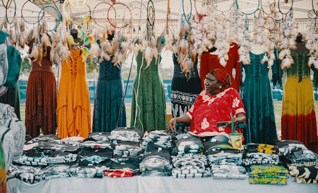 Black woman sits in chair at vendor booth, surrounded by colorful African dresses