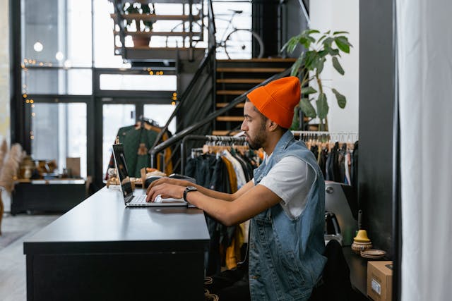 Light-skinned black man wearing blue jean vest and orange hat types on laptop near store counter