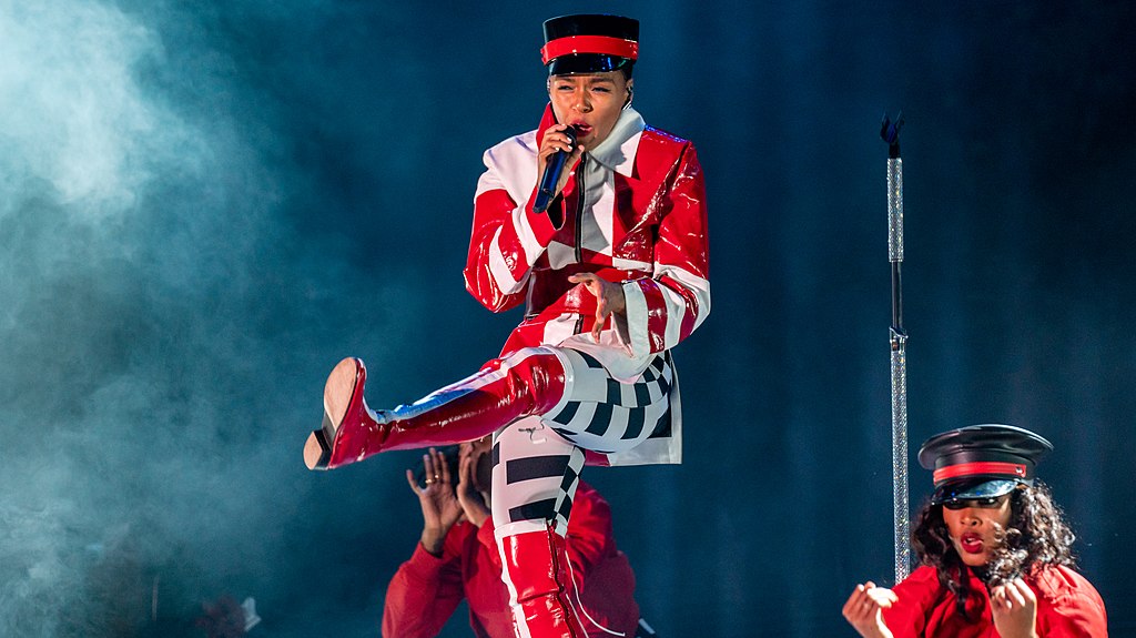 Janelle Monae wearing a red, white and black outfit while performing with her leg kicked out