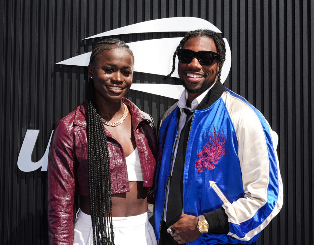 Olympian Junelle Bromfield, fiancee of Olympian Noah Lyles also pictured hrre, posing together and smiling, Bromfield in a burgundy leather jacket, white top and pants, Lyles in a blue, white and red satin bomber jacket wearing black sunglasses and a black tie